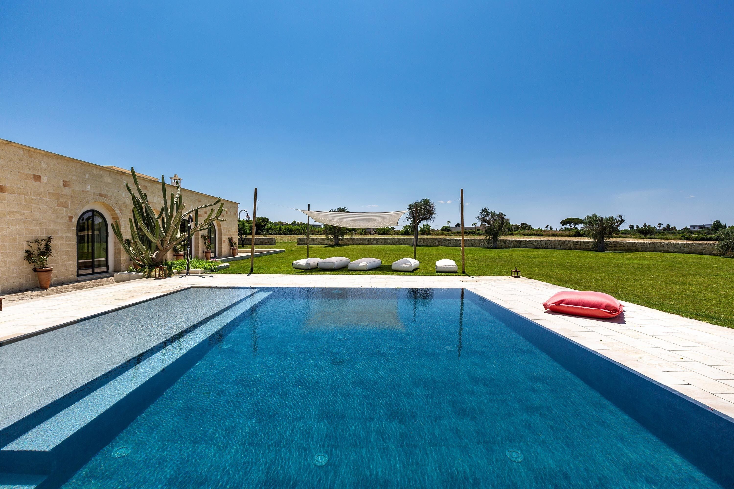 Una piscina con superficie d'acqua cristallina e azzurra, circondata da una zona pavimentata con piante in vaso e posti a sedere all'aperto.