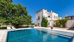 Une piscine rectangulaire à l’eau d’un bleu limpide, entourée d’une terrasse carrelée. On y trouve deux chaises longues et une petite table ronde près de la piscine. Un grand arbre est visible sur la gauche de la piscine.