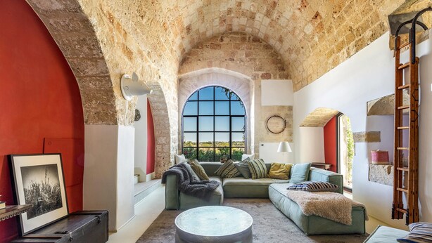 Un salon chaleureux avec une arche en pierre, une grande fenêtre, un canapé vert, une table basse ronde et une échelle appuyée contre le mur.