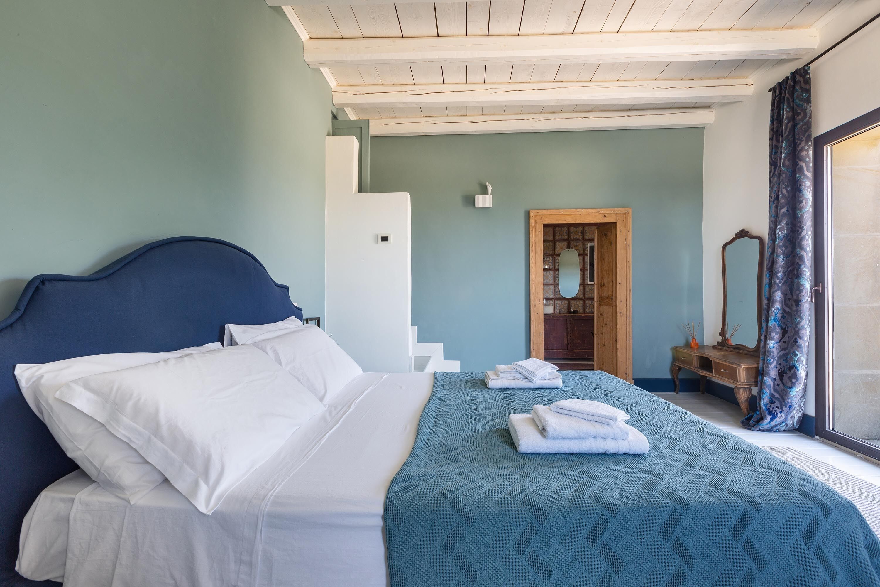 A neatly made bed with white linens and a teal blanket, a wooden nightstand with a mirror, and a view of a wooden door and a mirror in the background.