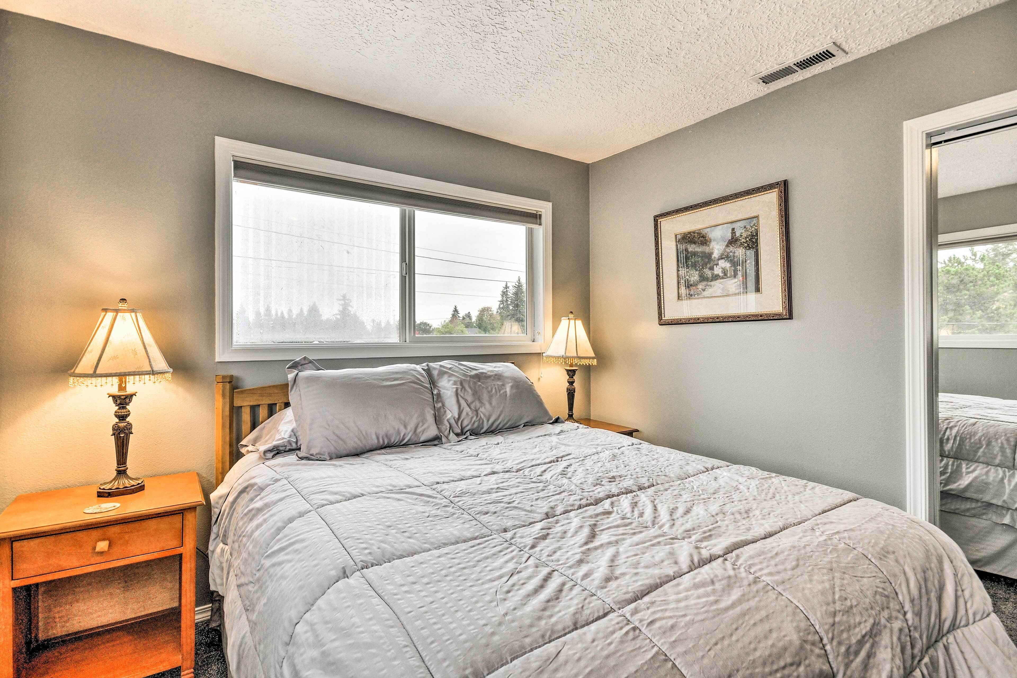 A bedroom with a large window, a bed with a quilted cover, two bedside lamps, a framed picture on the wall, and a wooden nightstand.