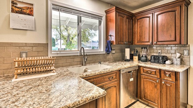 A kitchen with granite countertops, wooden cabinets, a built-in dishwasher, and a window with a view of trees. - Vancouver Home w/ Hot Tub < 15 Mi to Portland! (Vancouver)