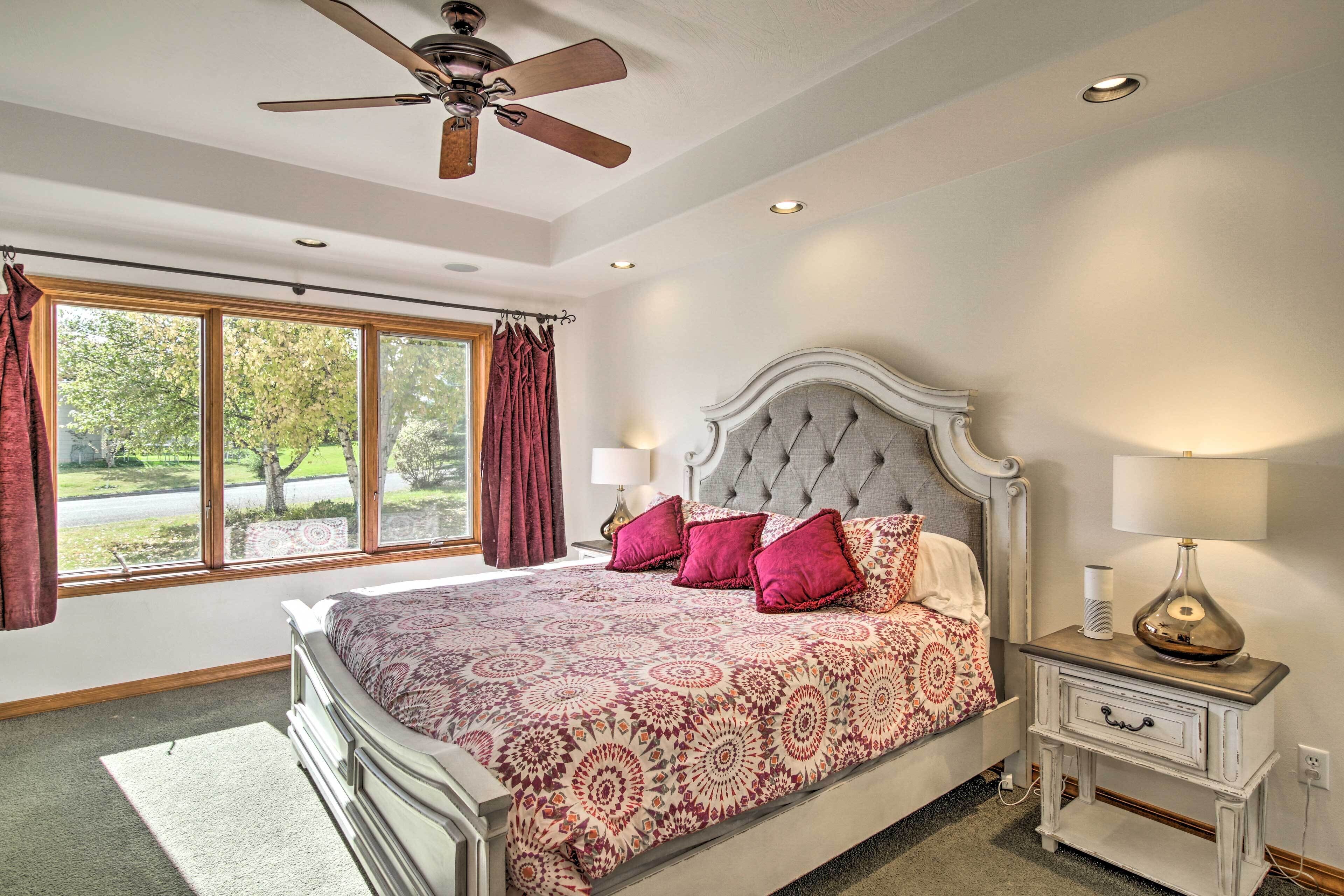 A bedroom with a large bed, a ceiling fan, a nightstand with a lamp, and a window with curtains.
