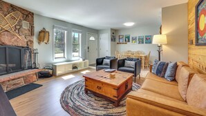 A living room with a stone fireplace, a wooden coffee table, a leather sofa, and a dining area with a table and chairs. - Quaint Winter Park Townhouse < 5 Mi to Ski! (Winter Park)