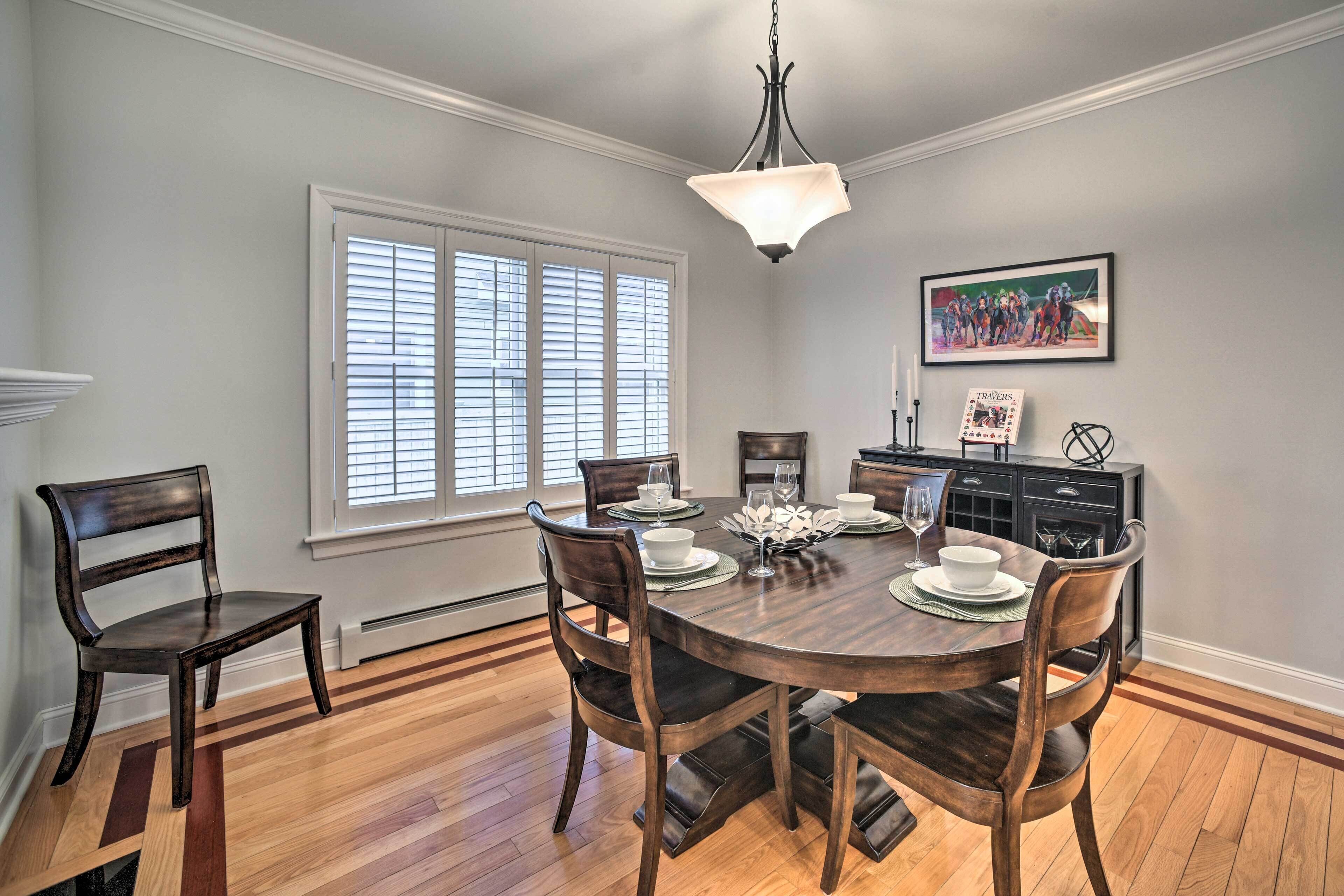 A dining area with a round table, six chairs, and a chandelier. There is a window with blinds, a painting on the wall, and a kitchen area in the background.