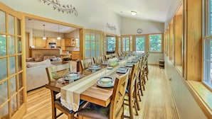 Ein langer Esstisch ist für eine Mahlzeit in einem geräumigen Zimmer mit Holzböden und großen Fenstern gedeckt.