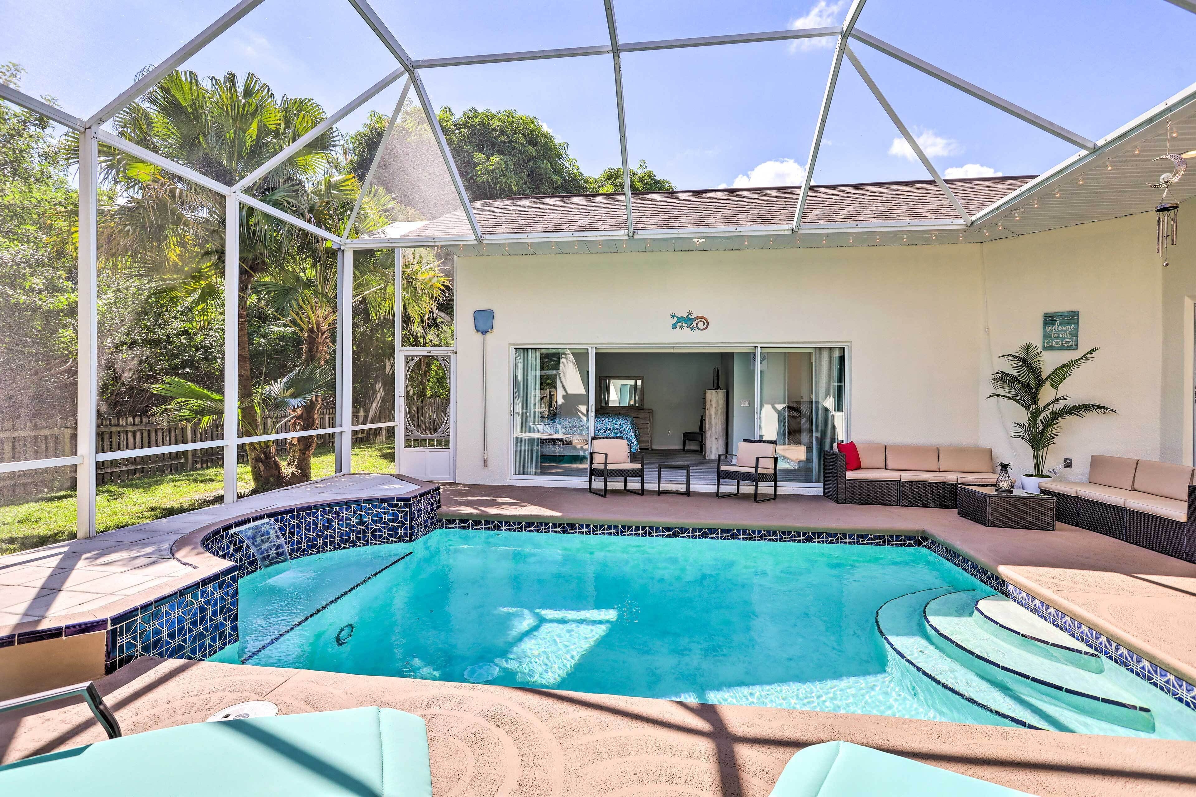 A covered swimming pool with a patio area, a seating area with a sofa and chairs, and a view of the surrounding greenery.