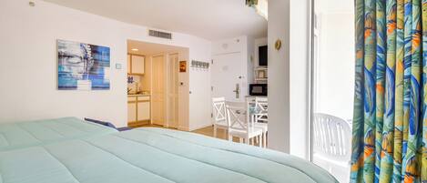 A bedroom with a bed, a dining area, and a kitchen visible through an open door.