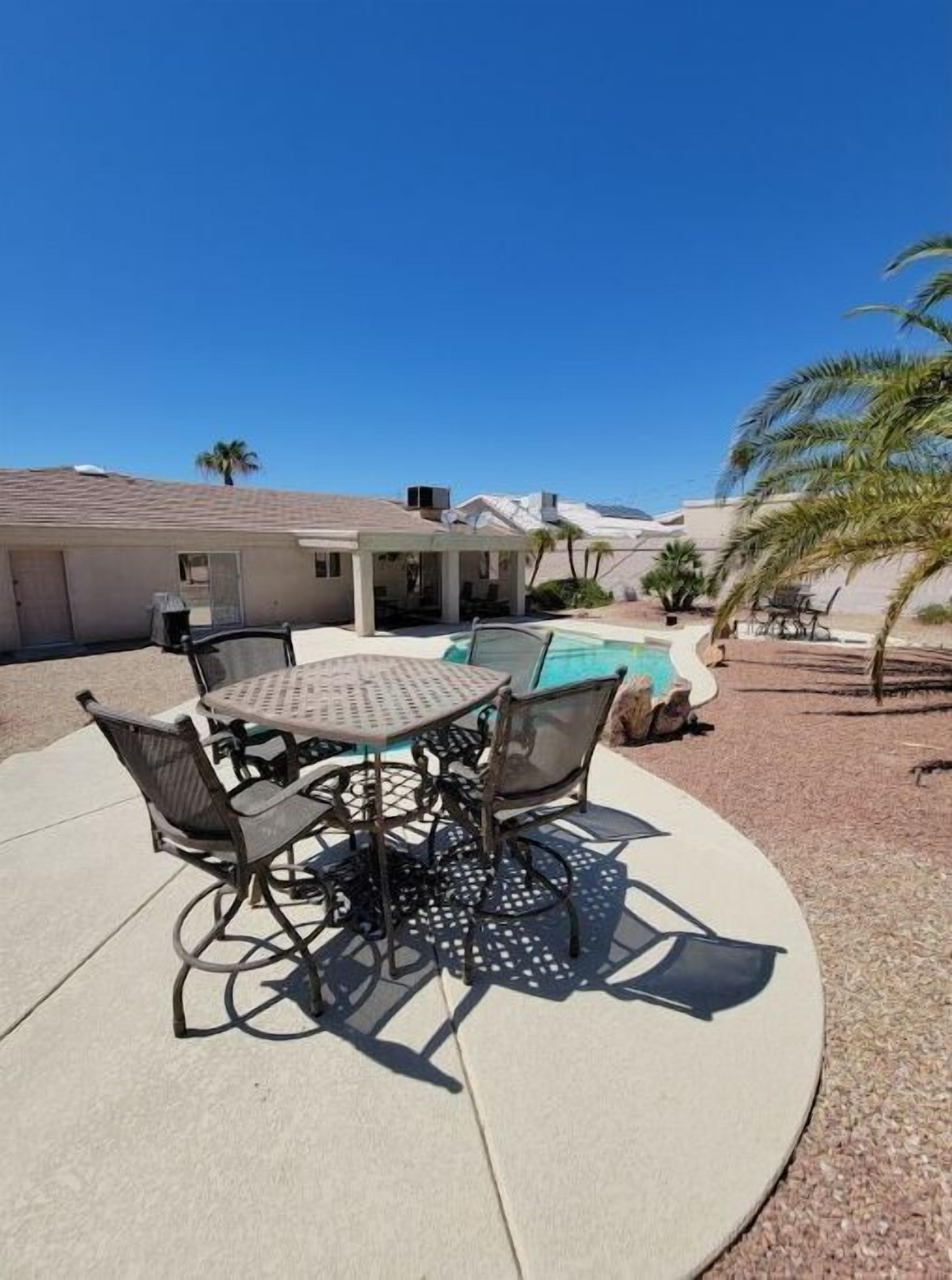 A patio with a round table and chairs, a pool, and a covered area with a view of mountains.