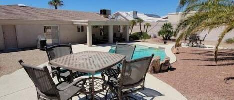 A patio with a round table and chairs, a pool, and a covered area with a view of mountains.