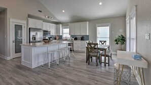A modern kitchen with white cabinetry, a central island with stools, a dining area with a table and chairs, and a small white table with a plant. - < 1 Mi to Sea Isle Beach: Home w/ Pool Access (Galveston)