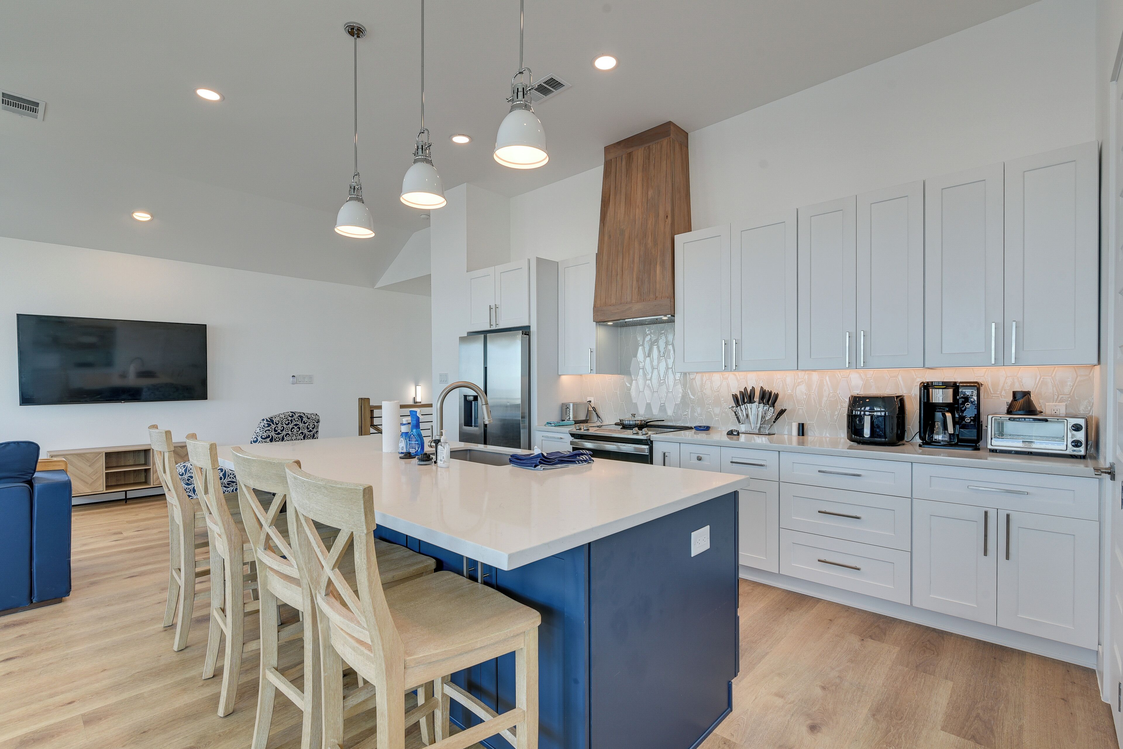 A modern kitchen with a central island, white cabinetry, and a large flat-screen TV mounted on the wall.