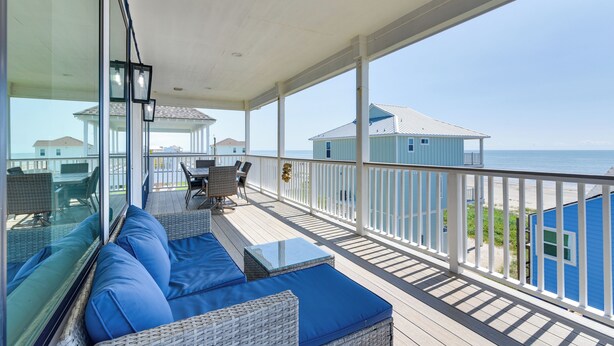A balcony with blue cushions, wicker furniture, and a view of the ocean and beach houses.