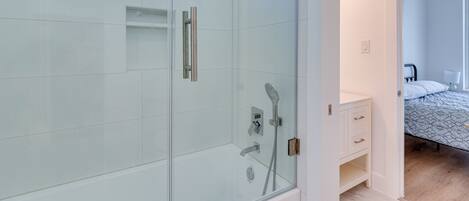 A modern bathroom with a glass shower enclosure, a white vanity, and a bed with patterned bedding.