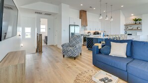 A modern living room with a blue sofa, a patterned armchair, and a kitchen area with white cabinets and a stainless steel refrigerator.