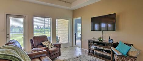 A spacious living room with a leather sofa, a coffee table, and a flat-screen TV mounted on the wall. There are large windows with blinds, a ceiling fan, and a patterned rug on the floor.