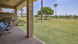 A covered patio with a grill and lounge chair, overlooking a well-maintained lawn and palm trees.