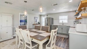 Un coin repas avec une table en bois et des chaises blanches, une cuisine équipée d’un réfrigérateur et d’un four à micro-ondes, et des luminaires suspendus.