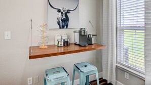 Un coin cuisine avec une étagère en bois où sont posés un appareil à café, des bocaux et un objet décoratif. On y trouve également deux tabourets et une fenêtre avec des stores.