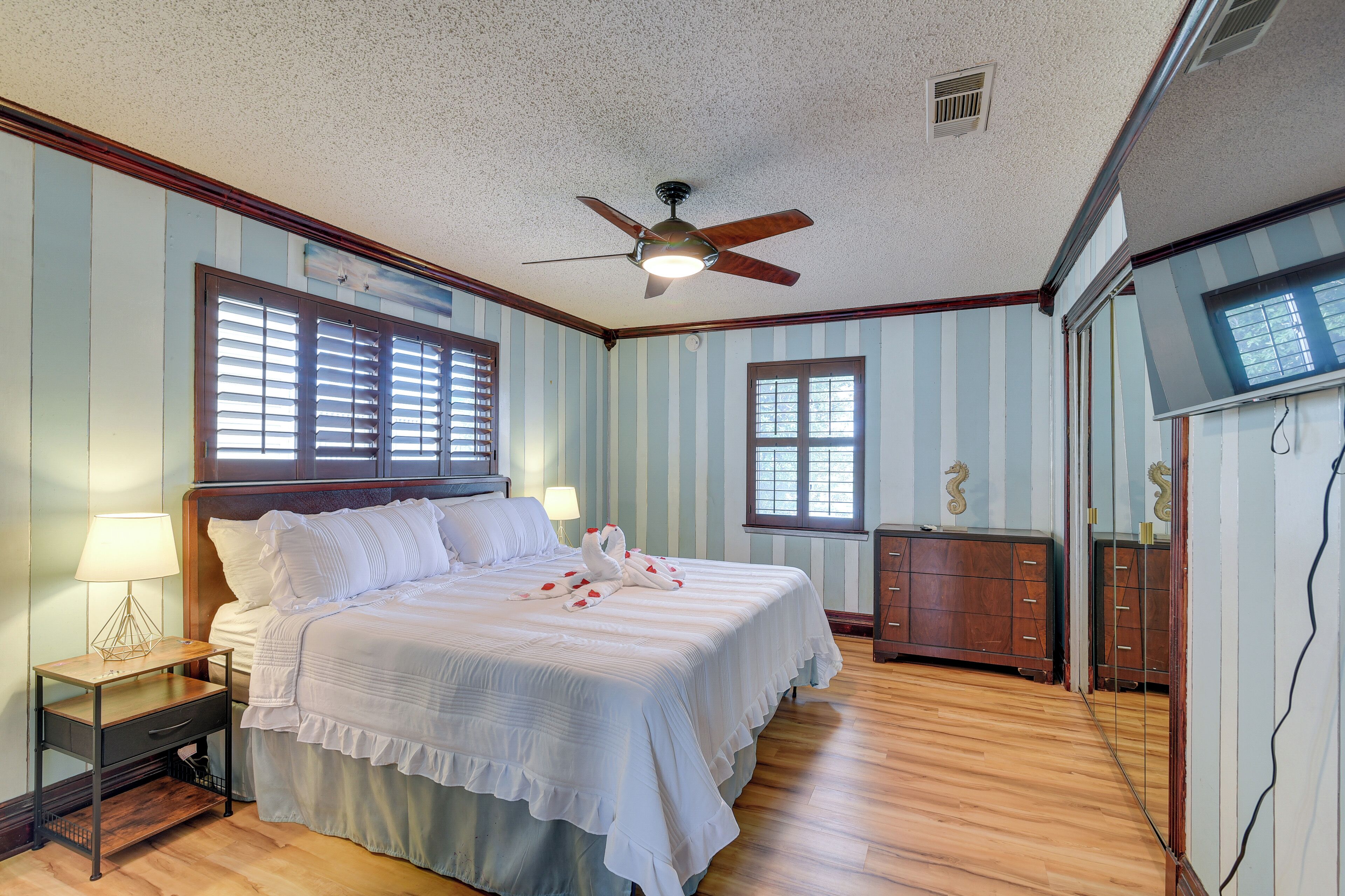 Un dormitorio con cama, mesita de noche, ventilador de techo y dos ventanas.