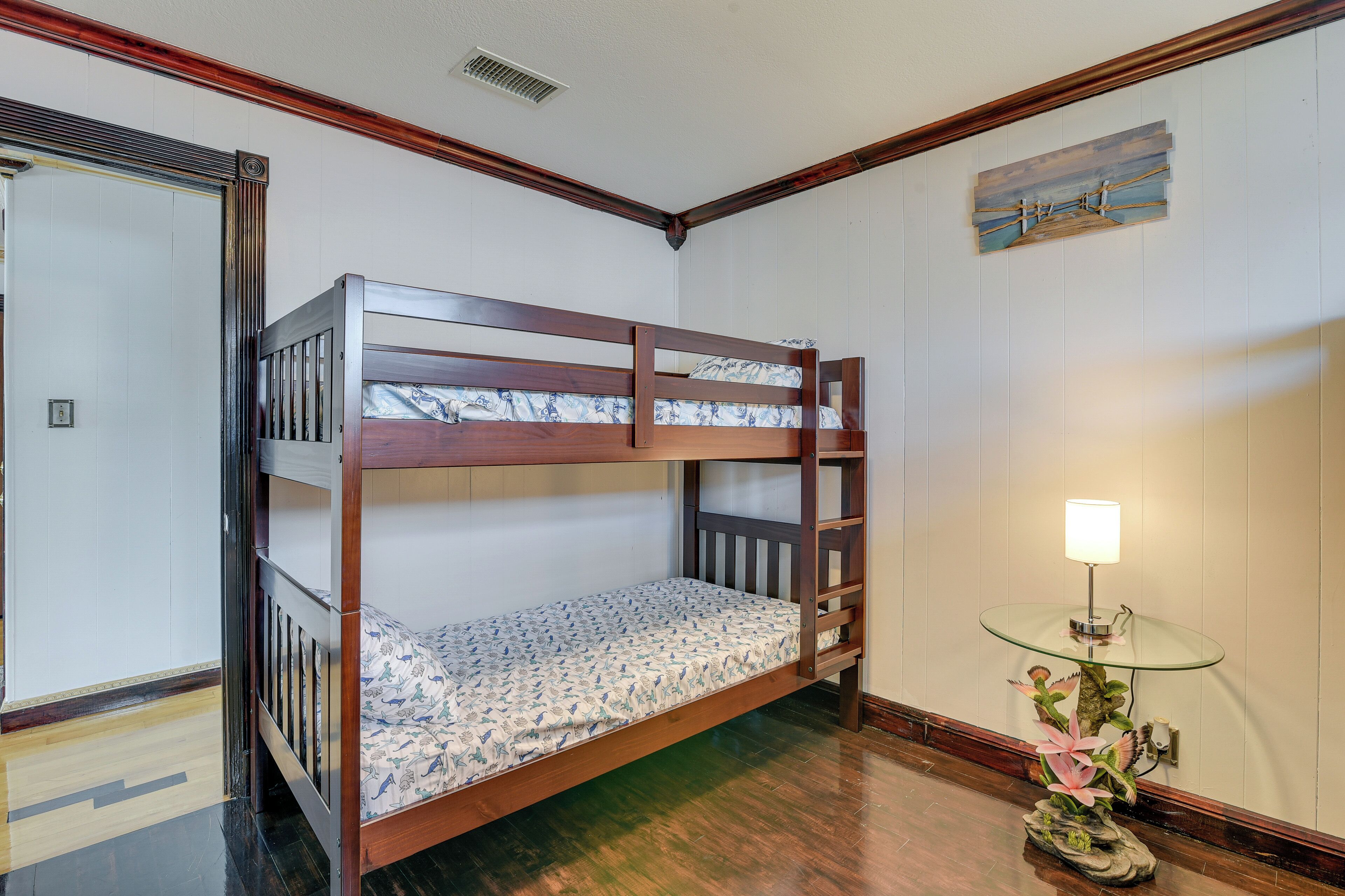A bunk bed in a room with wooden flooring, a small table with a lamp, and a decorative plant.