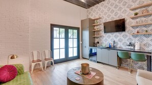 Un salon moderne avec un canapé vert, deux fauteuils blancs, une table basse ronde et un coin cuisine équipé d’un téléviseur, d’étagères et d’une fenêtre.