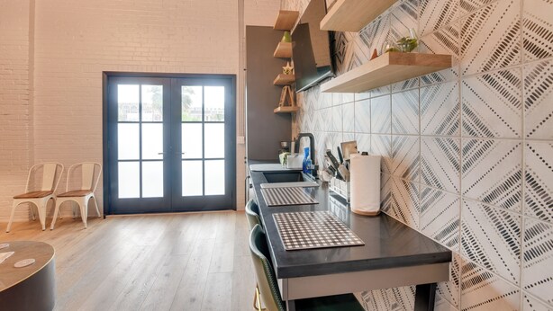 Une cuisine moderne avec un plan de travail noir, des murs en briques blanches et des carreaux décoratifs. On y trouve deux chaises blanches et une table en bois.