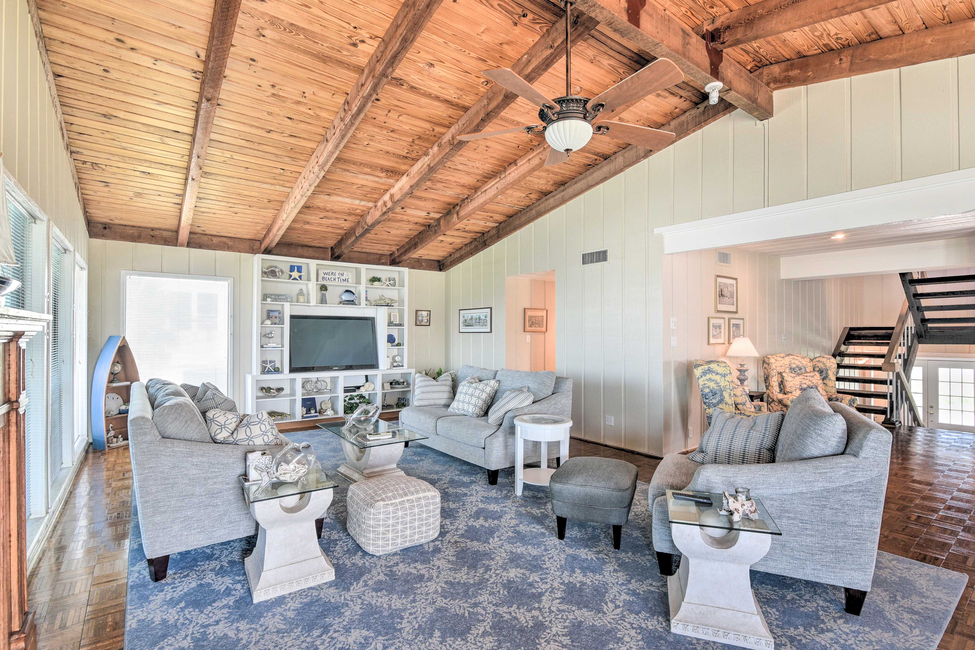 A spacious living room with a high ceiling and exposed beams, furnished with a grey sofa, ottomans, and a glass coffee table.
