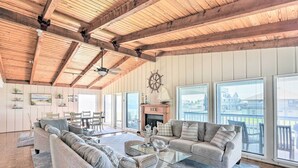 A spacious living room with a high ceiling and wooden beams, furnished with grey sofas and a glass coffee table, large windows offering a view of the outside.