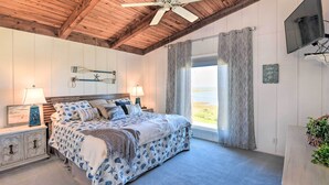 A bedroom with a bed, wooden ceiling, ceiling fan, window with curtains, and a TV mounted on the wall.