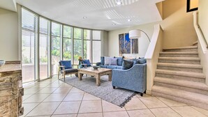 A spacious living room with a large window, a blue sofa, a wooden coffee table, and a staircase. - Bright & Airy Lahaina Condo - Walk to Beach! (Kapalua)