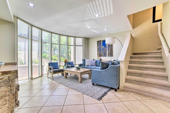 Un salón amplio con un gran ventanal, un sofá azul, una mesa de centro de madera y una escalera.