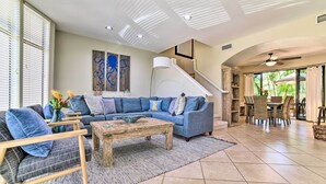 A spacious living room with a blue sectional sofa, a wooden coffee table, and a dining area with wicker chairs. - Bright & Airy Lahaina Condo - Walk to Beach! (Kapalua)