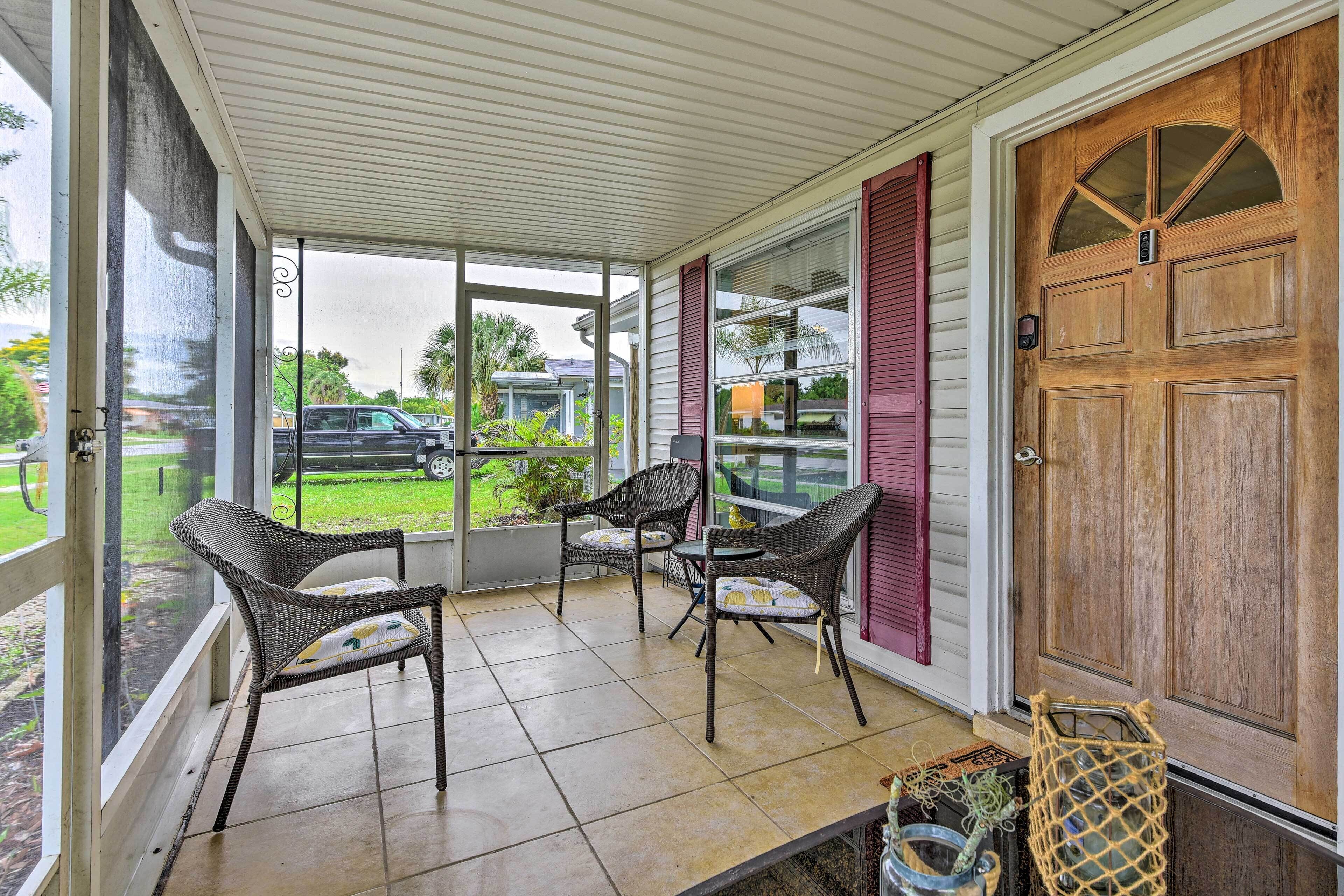 A covered porch with wicker chairs and a table, a wooden door, and a view of the outside.