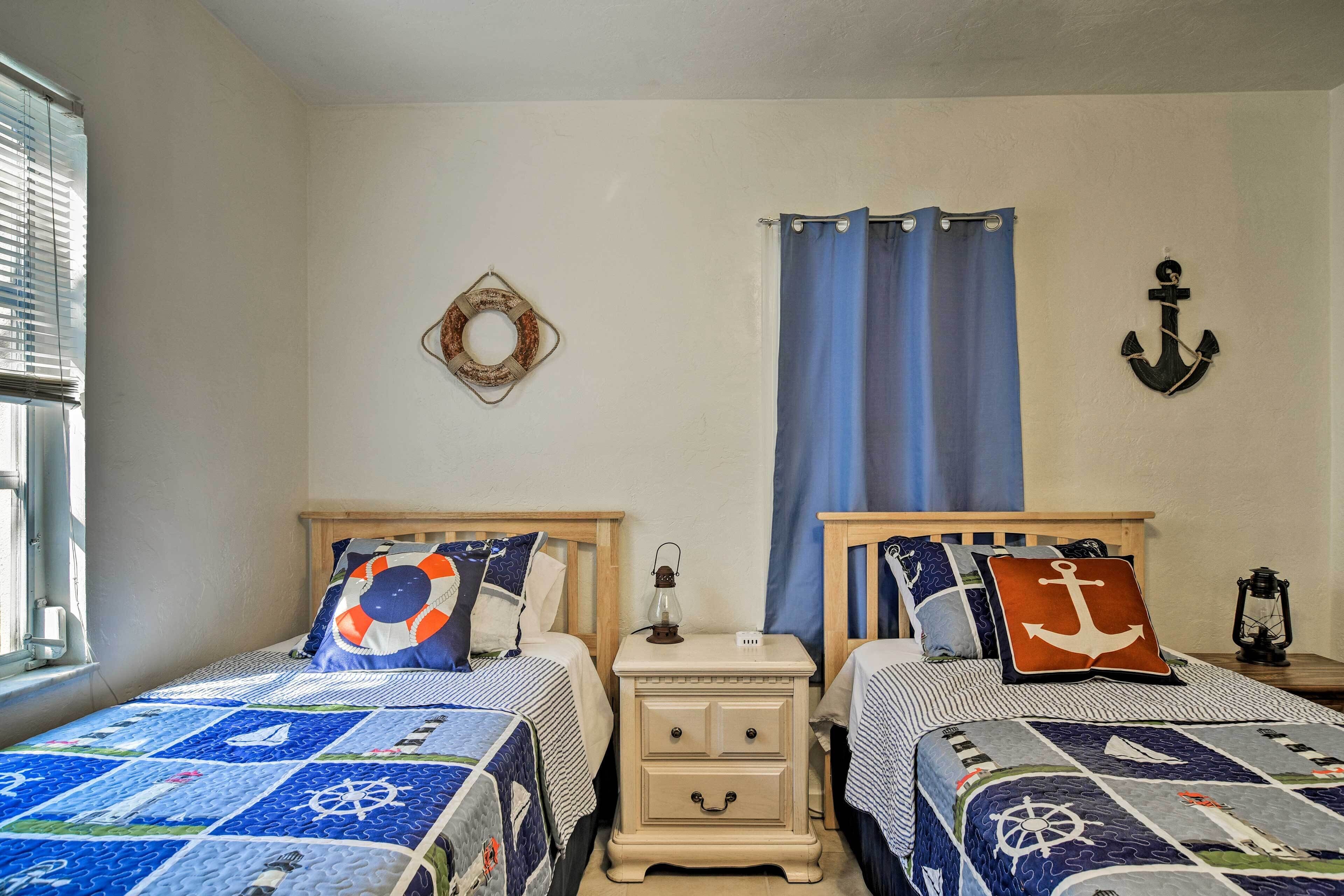 Un dormitorio con dos camas, ropa de cama con temática náutica, una mesita de noche con un farolillo y una ventana con persianas.