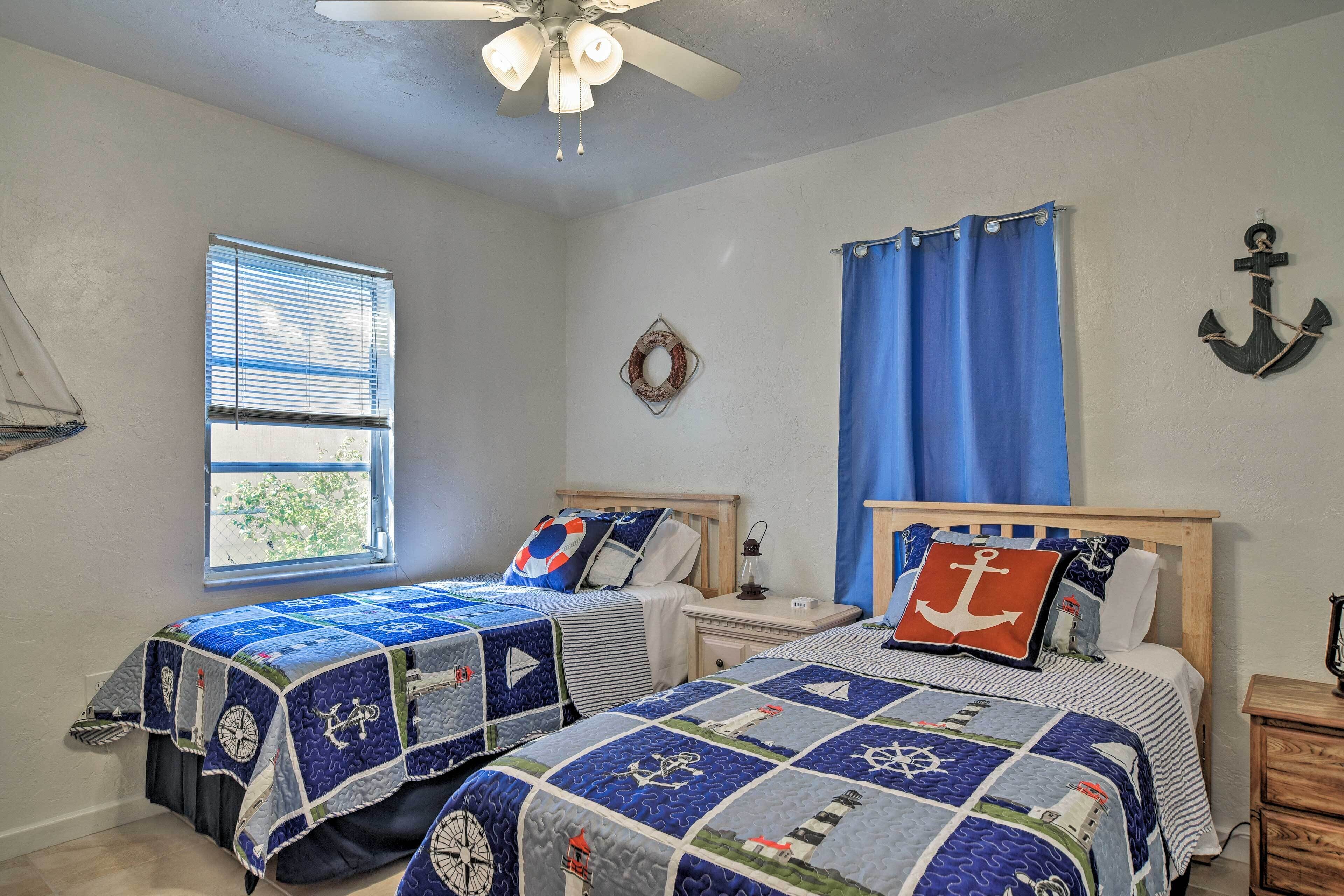 A bedroom with two beds, a window with blinds, a ceiling fan, and nautical-themed decor.