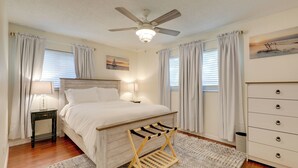 A bedroom with a bed, a ceiling fan, a nightstand with a lamp, a dresser, and a window with curtains.
