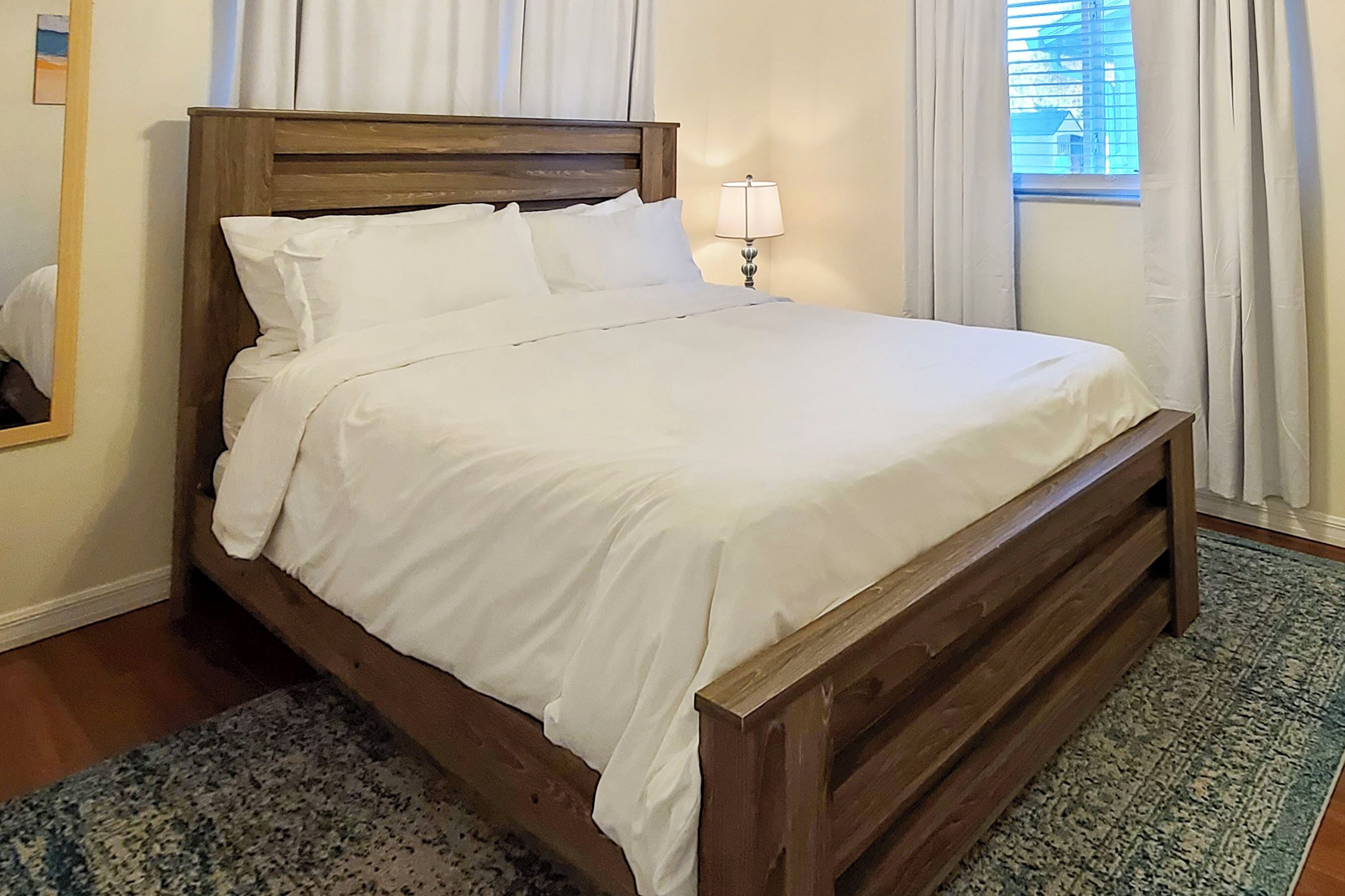 A neatly made bed with white linens in a room with wooden headboard, a patterned rug, and a lamp on a bedside table.
