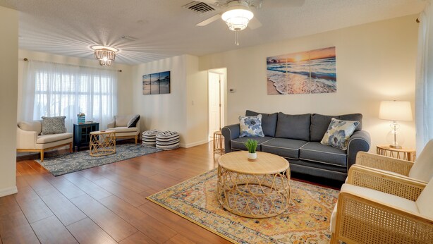 A spacious living room with a ceiling fan, a large window with sheer curtains, a grey sofa with blue and white pillows, a round wicker coffee table, and a patterned rug.