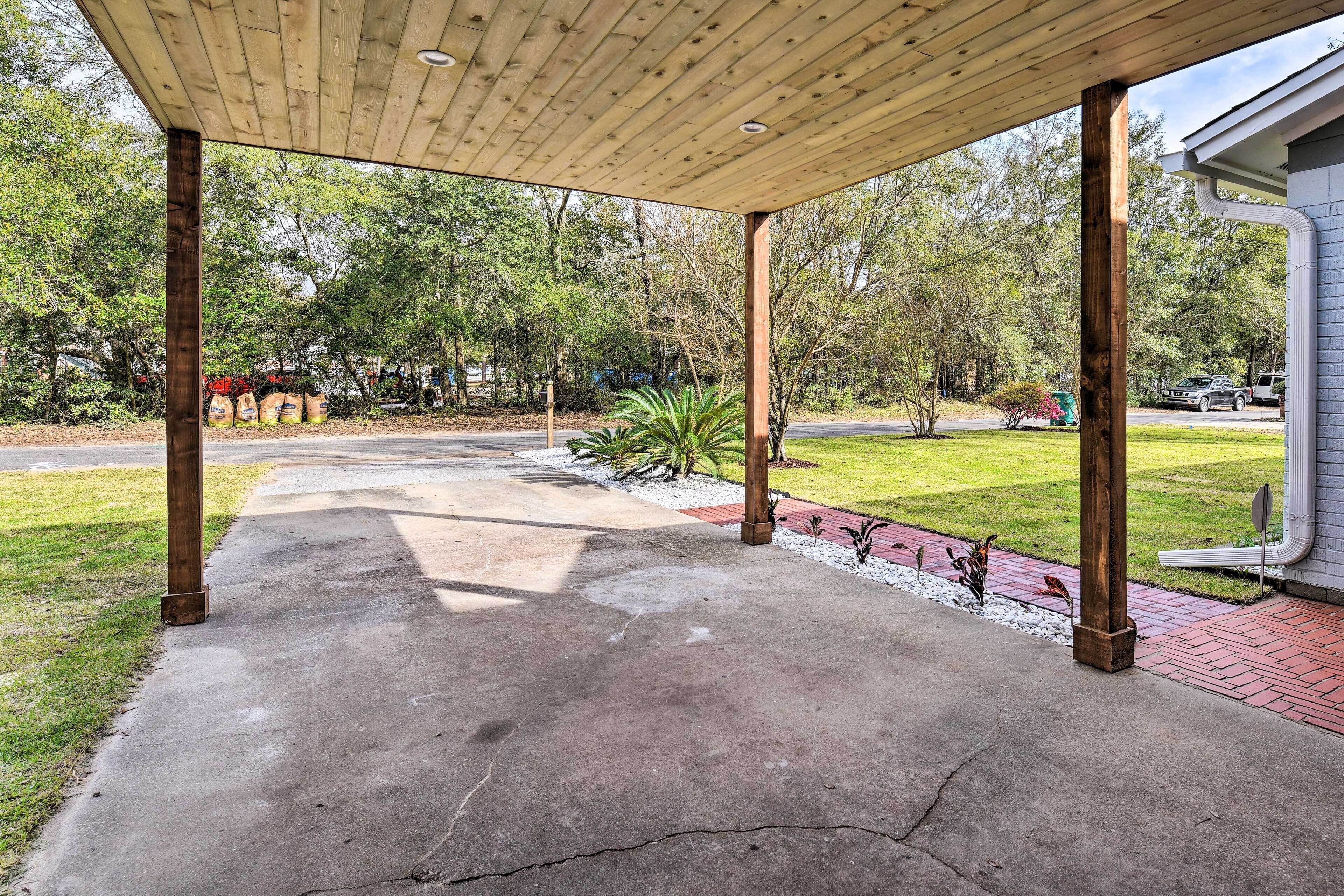 A covered patio area with a concrete floor, wooden pillars, and a view of a grassy yard with trees and a house in the background.