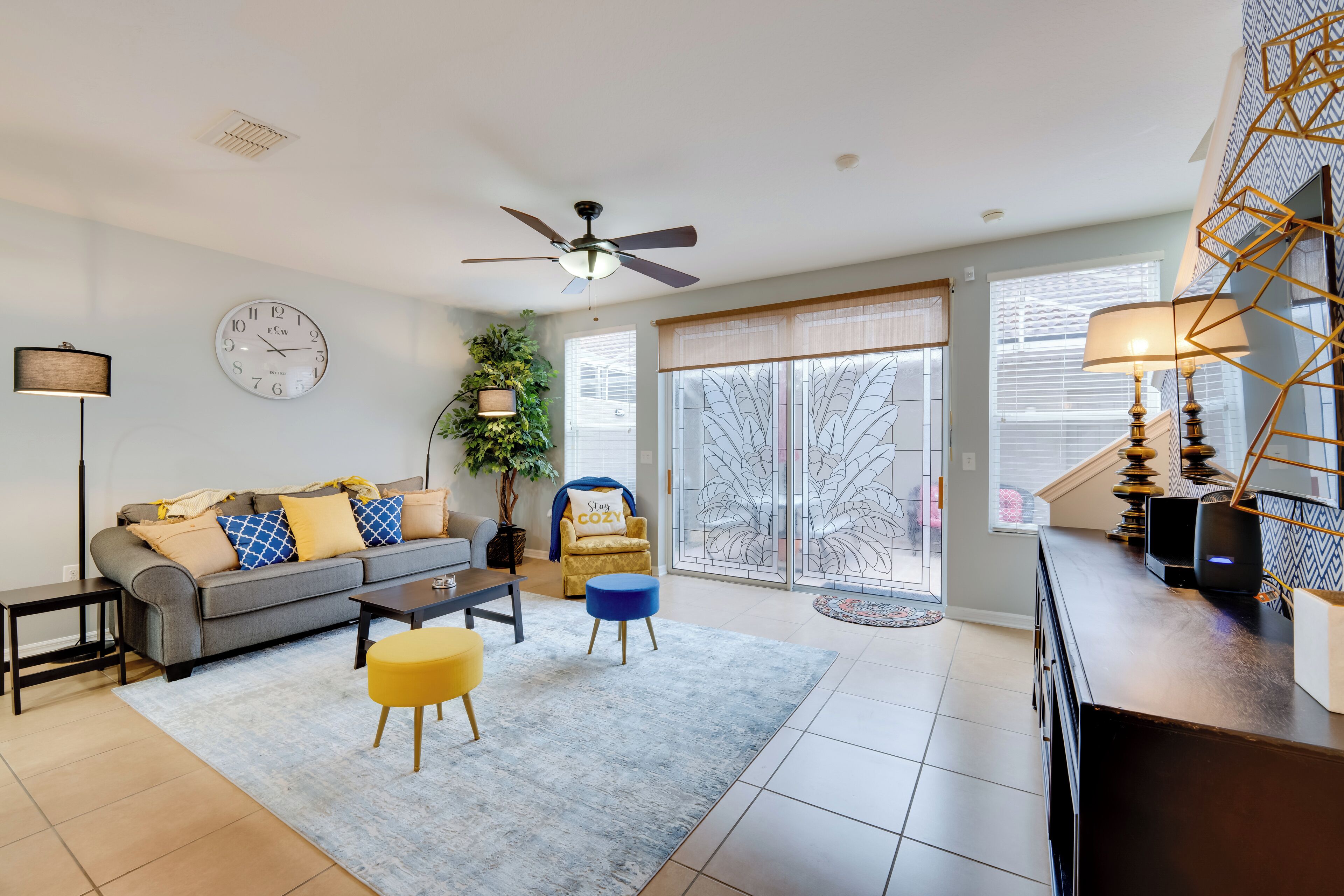 A modern living room with a sofa, coffee table, and ottoman, a ceiling fan, a clock, a plant, and a view through a glass door.