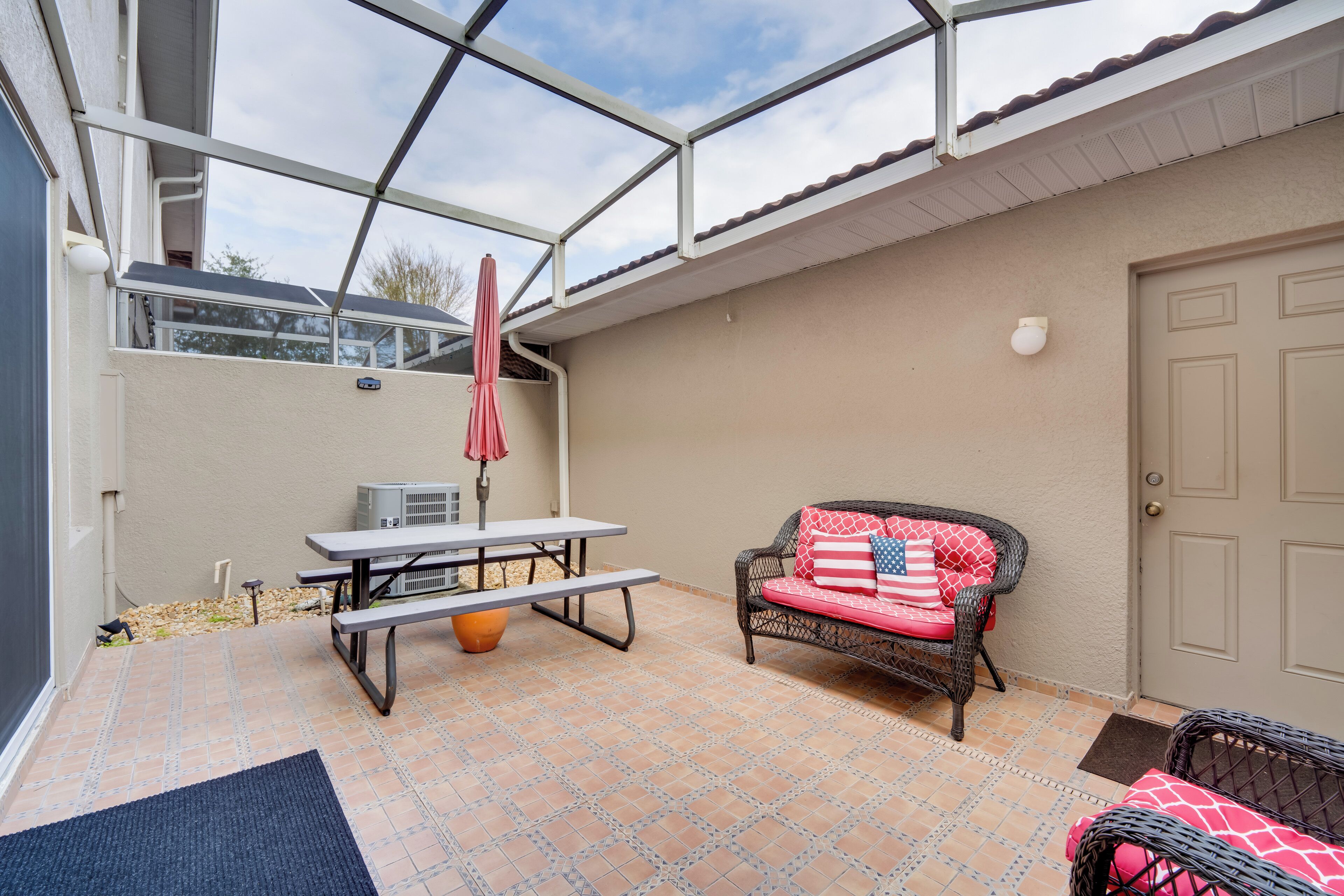 A covered patio with a picnic table, a wicker sofa with red cushions, and a door to the right.