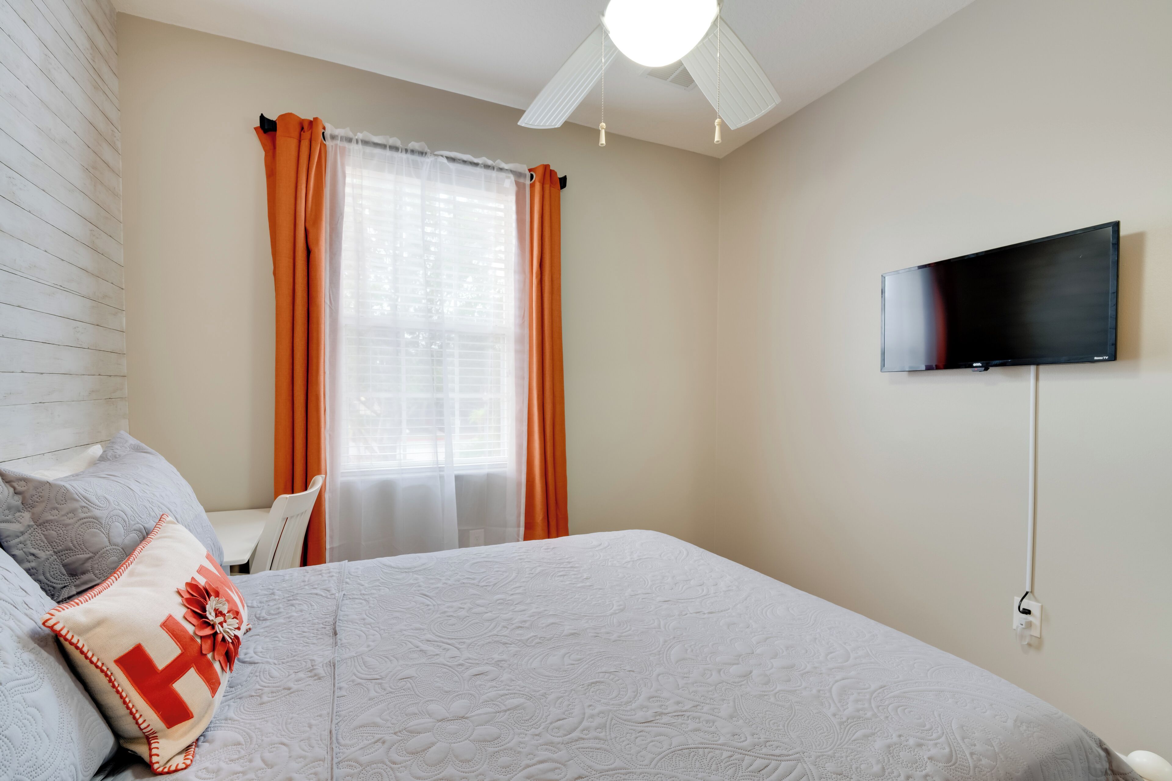 Una camera da letto ordinata con un letto, una televisione montata a parete e una finestra con tende.