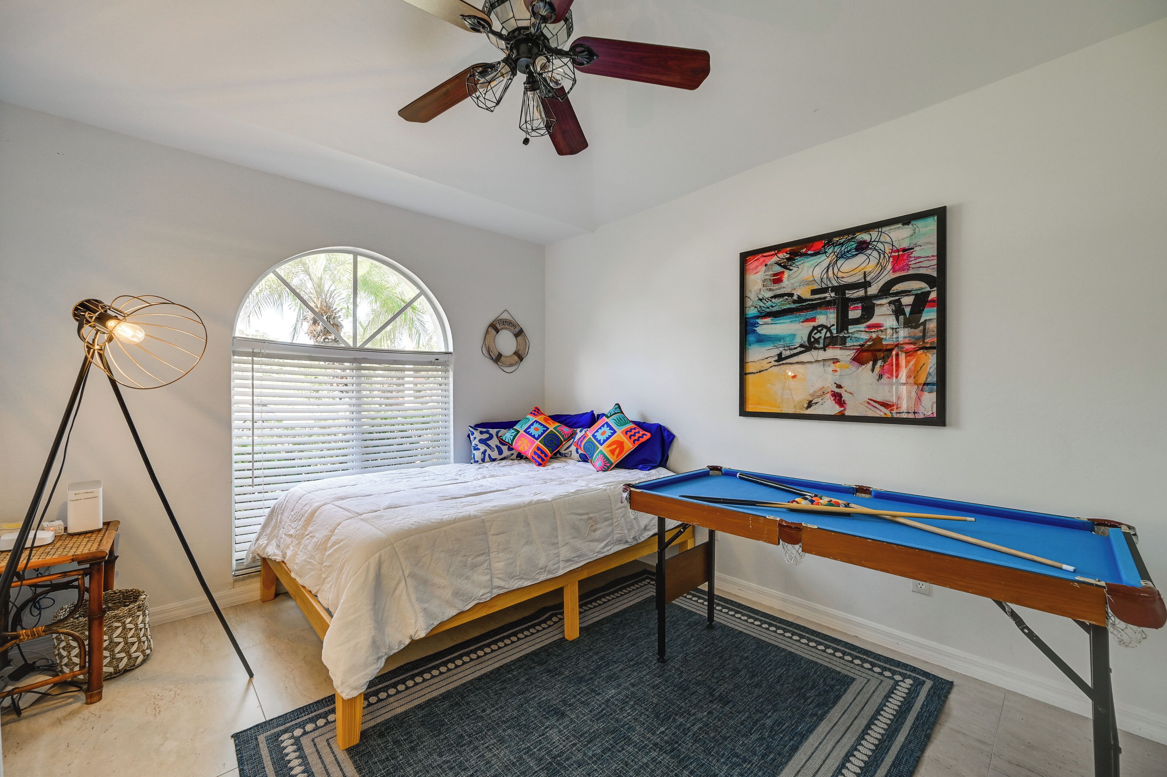 Une chambre à coucher avec un lit, une table de billard, un ventilateur de plafond, une fenêtre avec des stores et un tableau au mur.