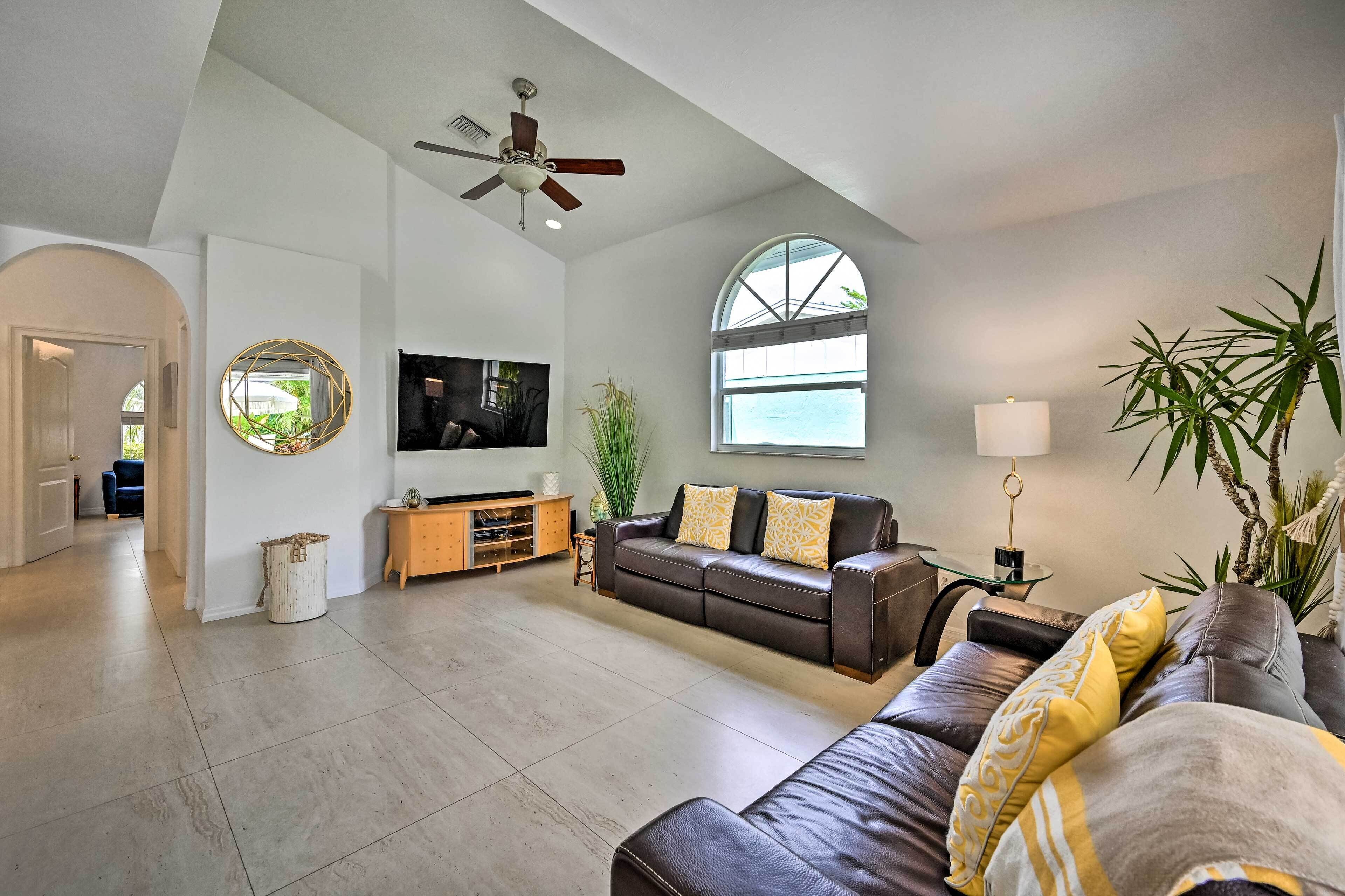 A spacious living room with a large window, a ceiling fan, a flat-screen TV, a leather sofa, and a wooden cabinet.