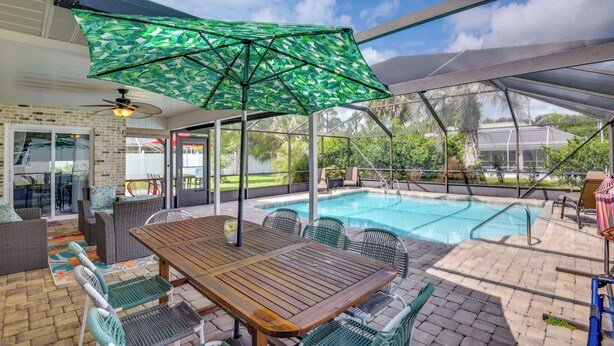 Un espace extérieur avec une piscine, une table et des chaises en bois, entouré d’une terrasse couverte agrémentée d’un parasol à motifs verts.
