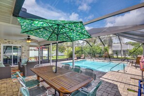 An outdoor pool area with a wooden table and chairs, surrounded by a covered patio with a green patterned umbrella. - Chic Palm Coast Escape: Lanai & Heated Pool! (Palm Coast)