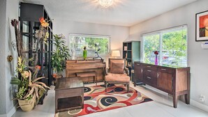 A modern living room with a glass coffee table, a brown armchair, a wooden cabinet, and a large window with a view of greenery. - Shady & Eclectic Fort Lauderdale Dwelling w/ Yard! (Fort Lauderdale)