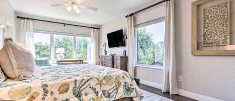 A bedroom with a bed, a ceiling fan, a TV, and large windows.
