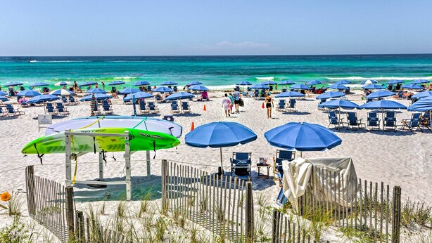 Une plage avec de nombreux parasols et des chaises longues, une clôture en bois et un porte-planches de surf.